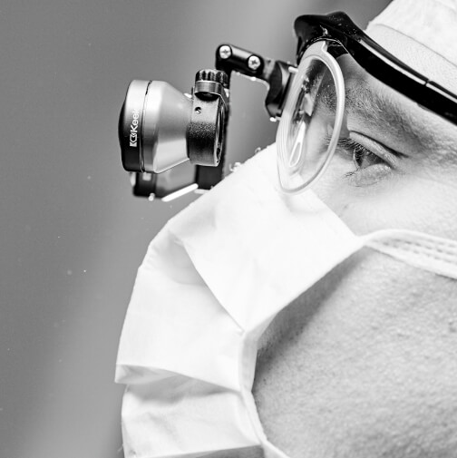 Eine Person, die eine Lupenbrille und eine chirurgische Maske trägt und von der Seite zusehen ist